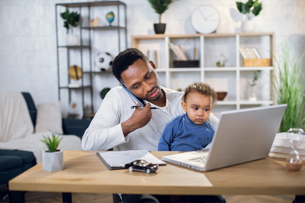 African businessman holding his cute son on knees while sitting at table with modern laptop and talking on mobile. Multitasking person at home.