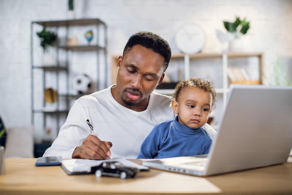 Focused young man working on laptop and writing on clipboard while sitting at table with baby boy on knees. African father taking care of his son and working from home.