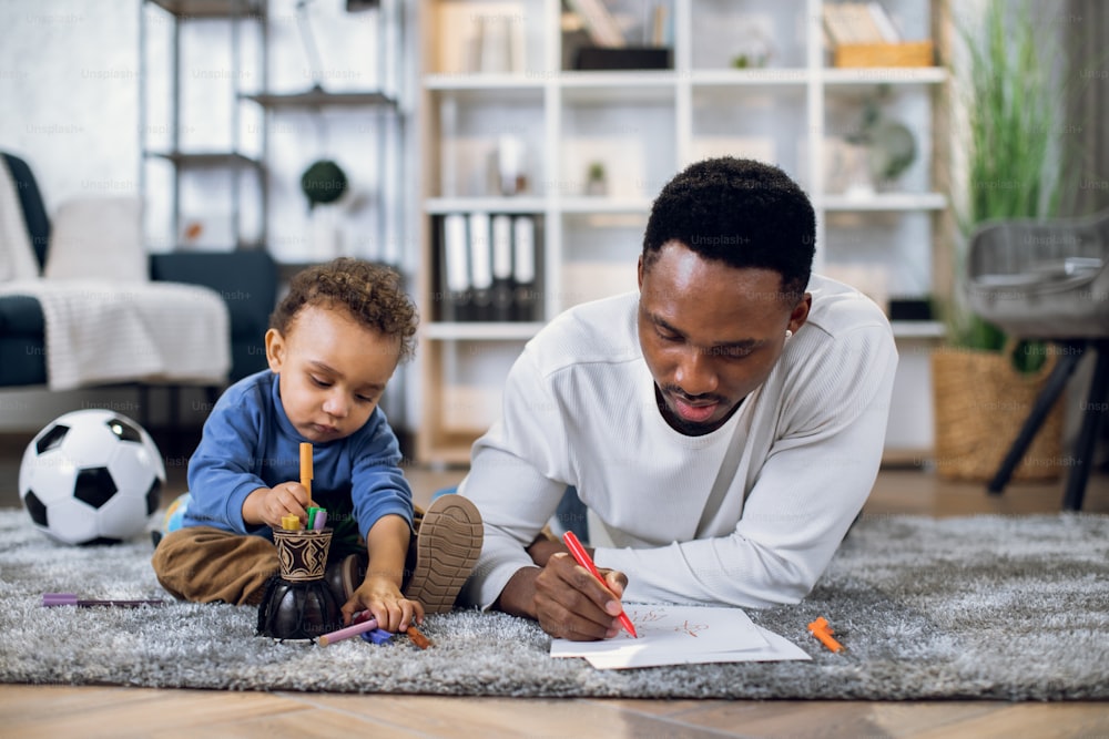 Liebevoller afroamerikanischer Vater und süßer Sohn, der zusammen auf dem Boden liegt und mit bunten Bleistiften im Album zeichnet. Familienzeit zu Hause. Konzept des Miteinanders.