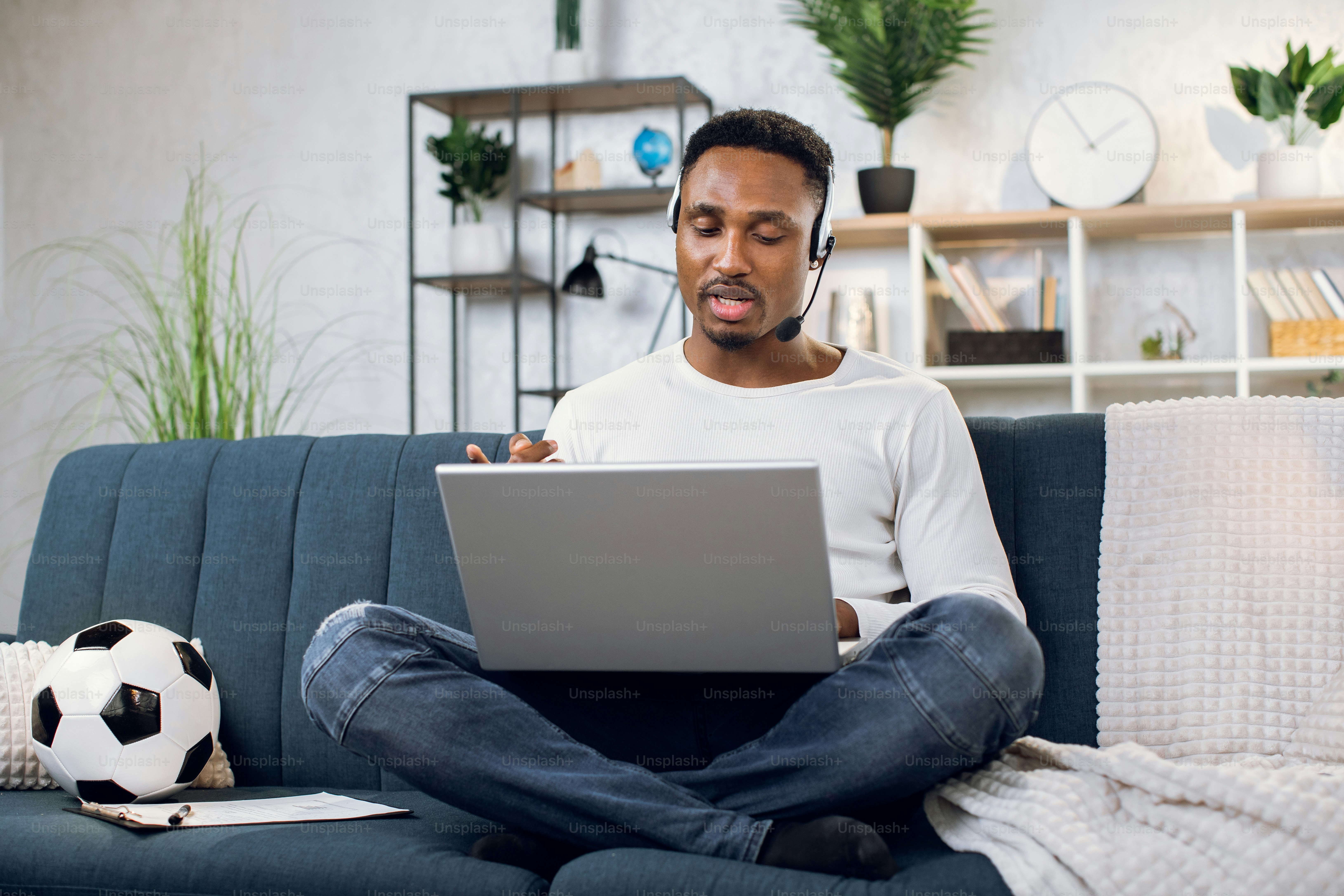 Casual african man in headset having video chat on modern laptop while sitting on cozy sofa. Young handsome guy working remotely while staying at home.