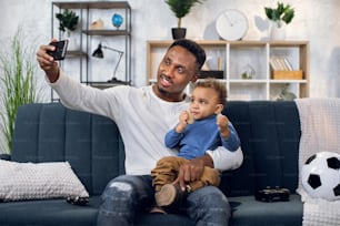 Happy afro american father and his cute son sitting together on couch and taking selfie on modern smartphone. Family time at home. Technology and people concept.