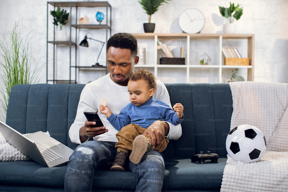 Hübscher afrikanischer Mann in legerem Outfit, der einen kleinen Jungen auf den Knien hält und ein modernes Smartphone benutzt. Junger Vater und Sohn spielen zusammen auf der Couch zu Hause.
