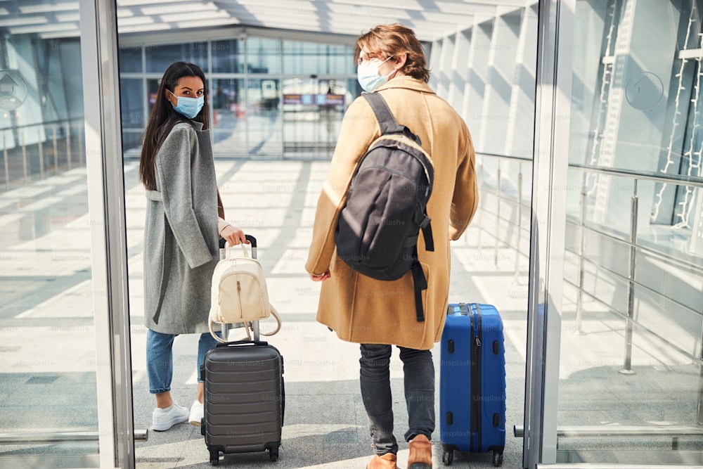 Elegant lady and gentleman wearing protective face masks while carrying travel suitcases at airport