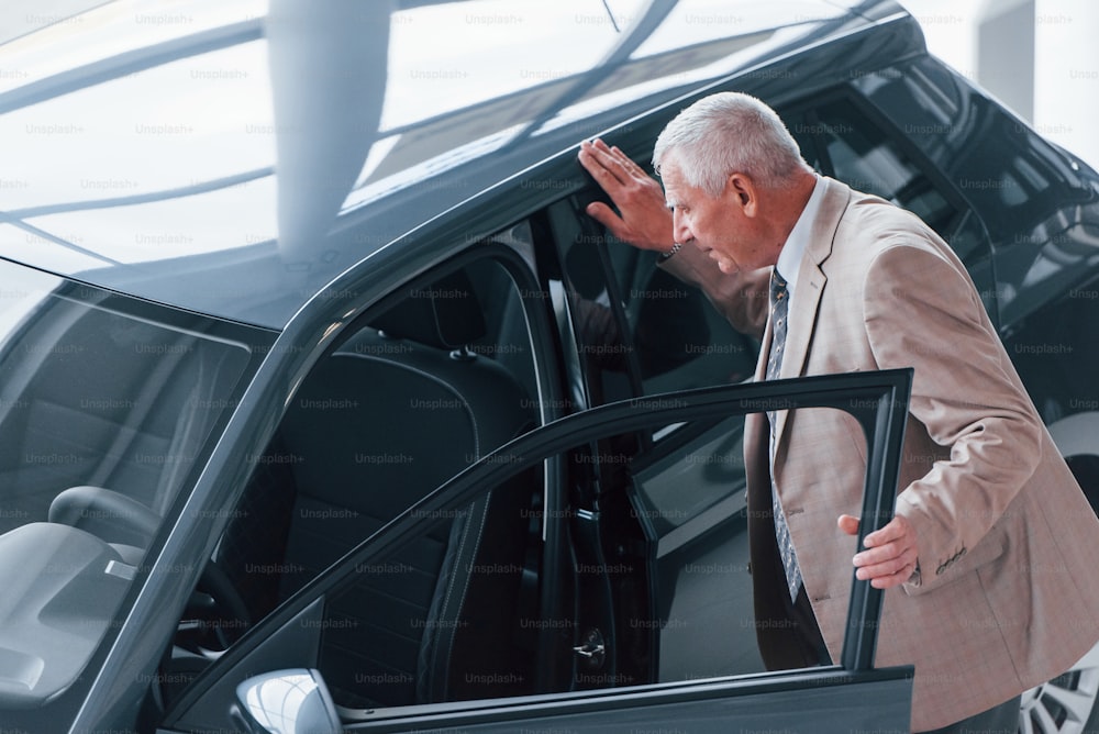 Aged man in formal wear walking in the automobile salon and looking for the car.