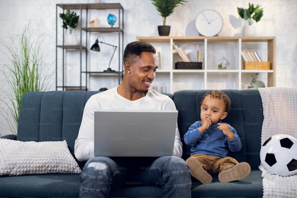 Young afro american father sitting with his cute son on comfy couch and working on wireless laptop. Male freelancer using portable computer for remote work at home.