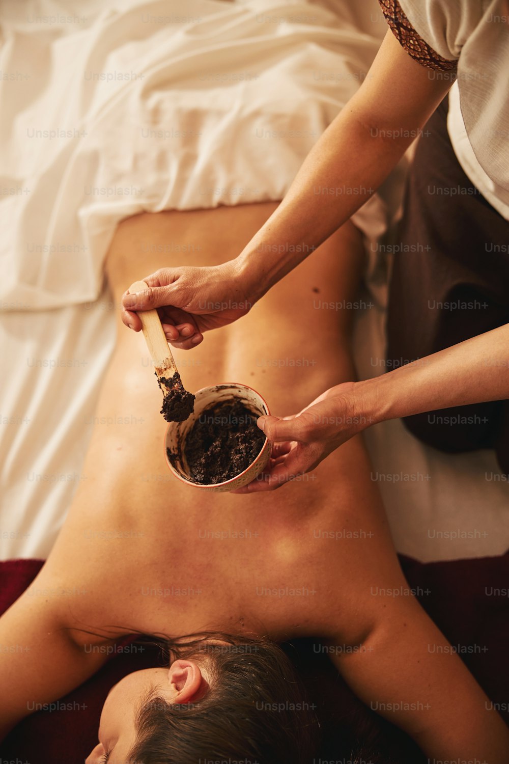 Woman without clothes on her back lying on bed while spa specialist keeping bowl with mud for back treatment