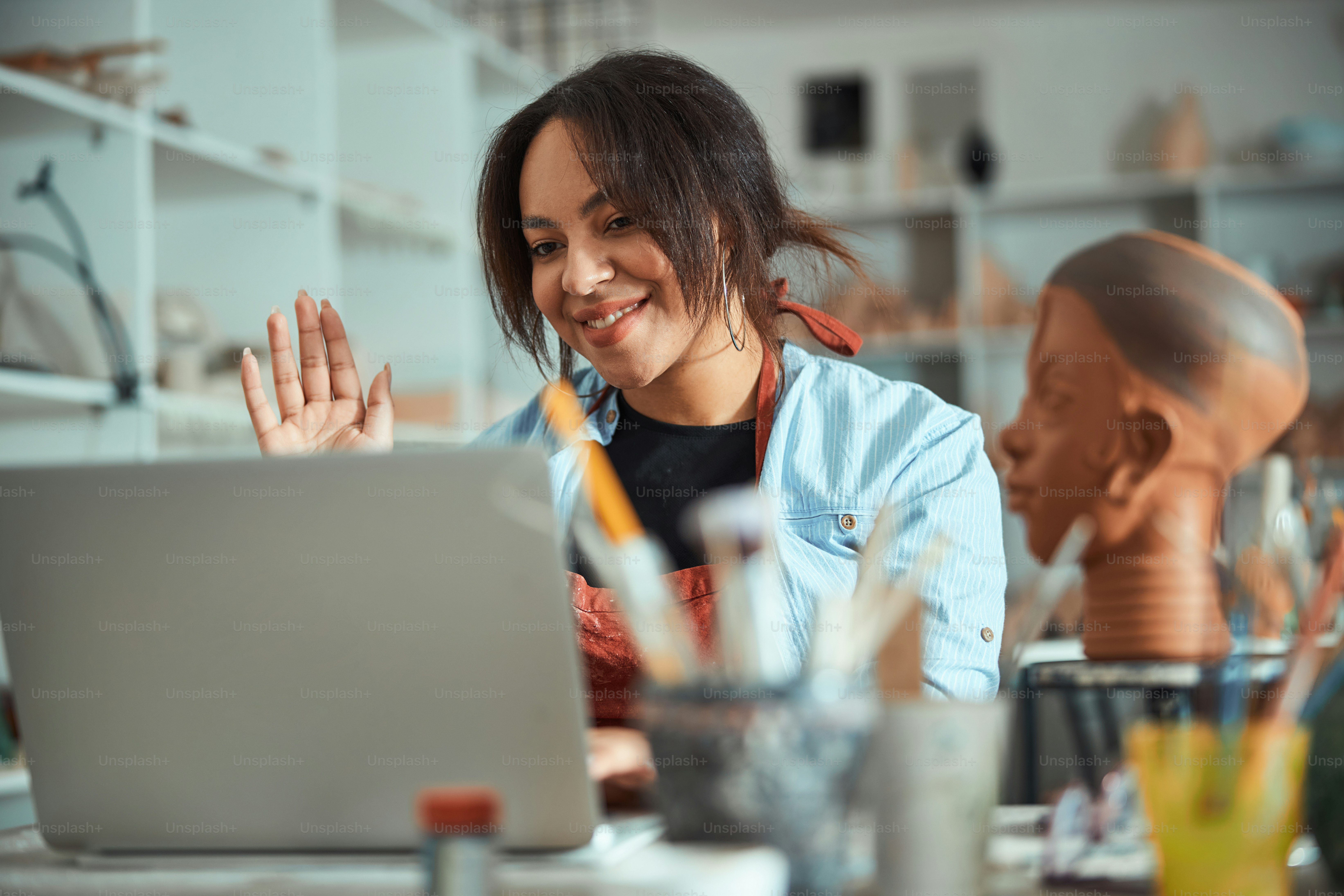 Charming young woman ceramic artist doing hello gesture and smiling while talking with friend through video call