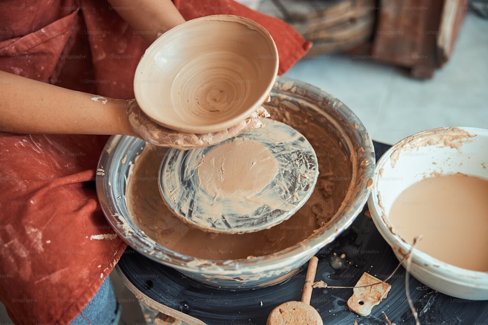 Gros plan d’une jeune potière avec un bol d’argile dans ses mains assise près d’un tour de potier dans un atelier