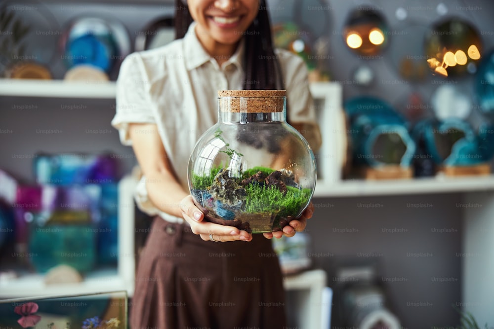 Photo recadrée d’une femme avec des tresses tenant un terrarium de plantes en bocal rond fermé devant la caméra