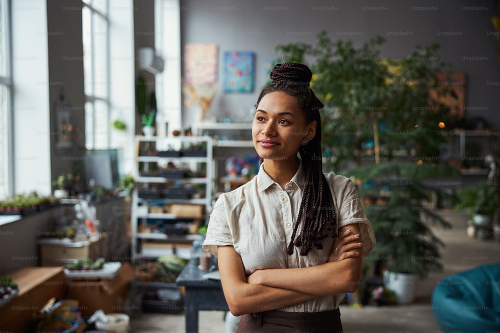 Waist-up portrait of a calm pleased attractive lady florist with cornrows standing in a workshop