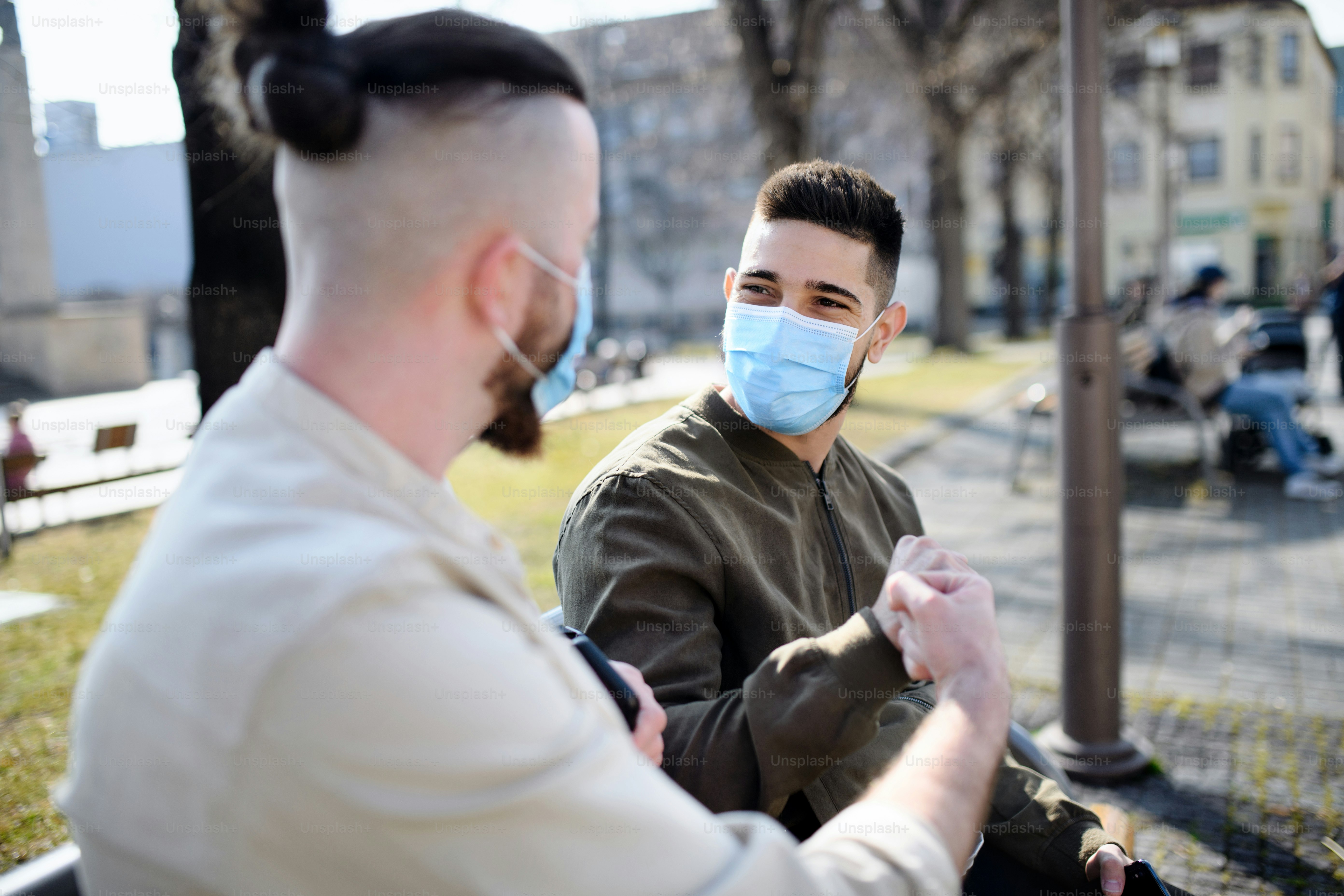 Cheerful men friends outdoors on street in town sitting and greeting, coronavirus and new normal concept.