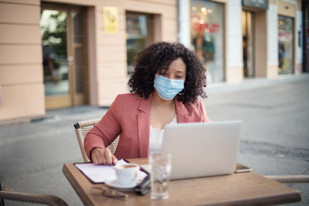 Frau trägt eine schützende Gesichtsmaske und benutzt einen Laptop im Café. Geschäftsfrau mit Schutzmaske sitzt im Café und arbeitet am Laptop.