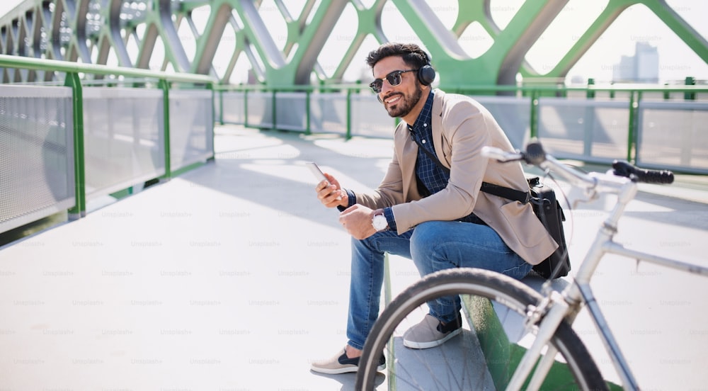 Portrait of young business man commuter with bicycle going to work outdoors in city, using smartphone.
