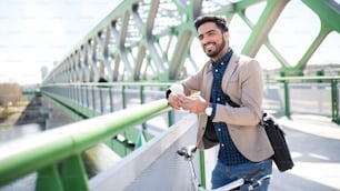 Ritratto di giovane pendolare uomo d'affari con bicicletta che va a lavorare all'aperto in città, usando lo smartphone sul ponte.