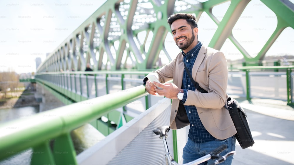 Portrait of young business man commuter with bicycle going to work outdoors in city, using smartphone on bridge.