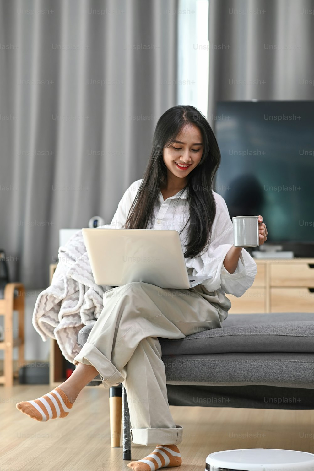 Happy woman using laptop on sofa and robotic vacuum cleaner cleaning in living room.