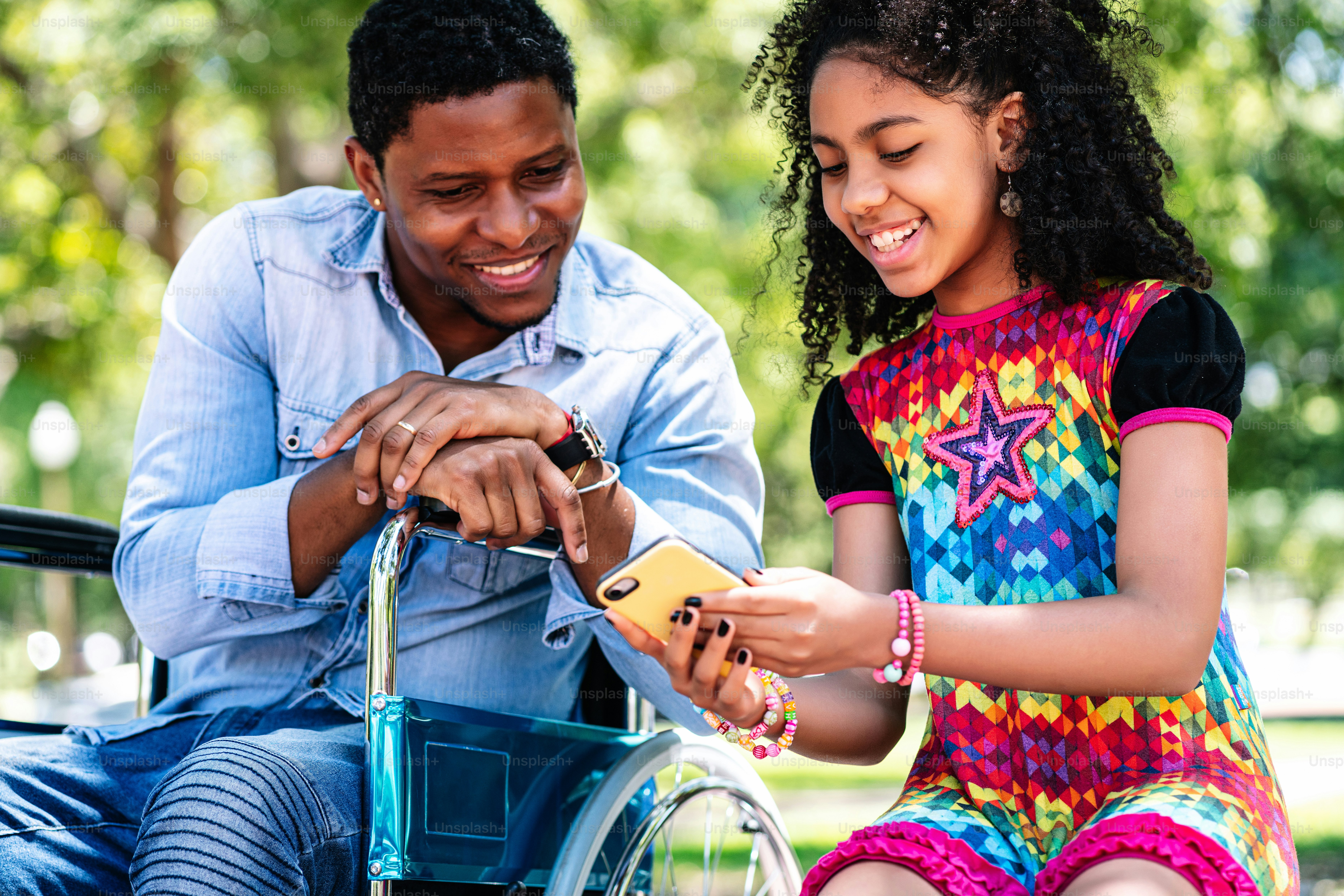 Un hombre afroamericano en silla de ruedas usando un teléfono móvil con su hija mientras disfrutan de un día juntos en el parque.