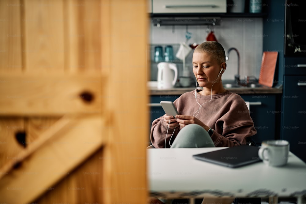 Pretty senior woman sitting in the kitchen at home and using phone for social media, listening to the music or video call.