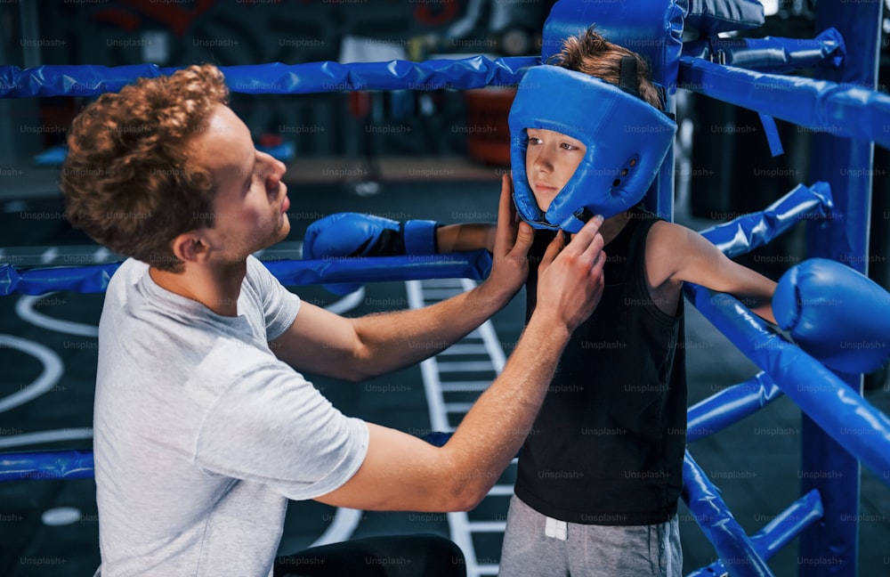 Young boxing coach is helping little boy in protective wear on the ring between the rounds.