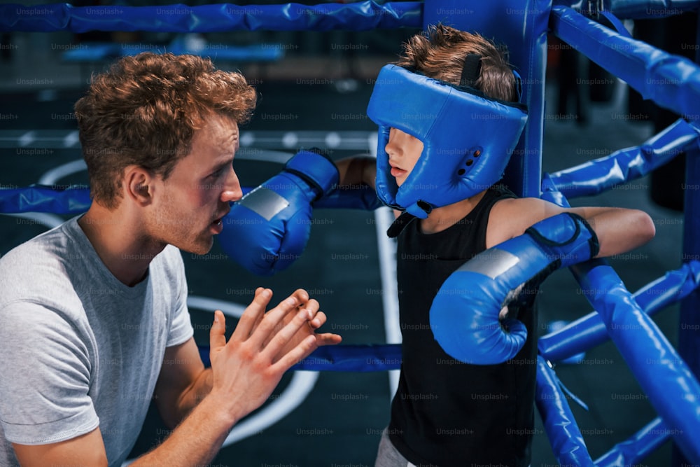Jovem treinador de boxe está ajudando garotinho em trajes de proteção no ringue entre os rounds.