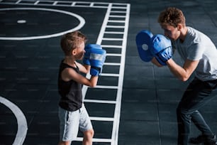 Young trainer teaches kid boxing sport in the gym.