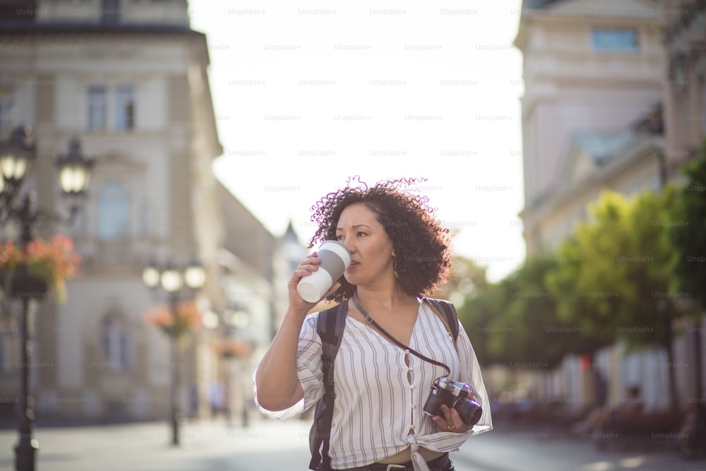 In search of the perfect picture. Woman with camera standing on street and drinking coffee.