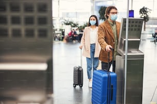 Male traveler in protective face mask checking luggage weight while woman standing in line at safe distance