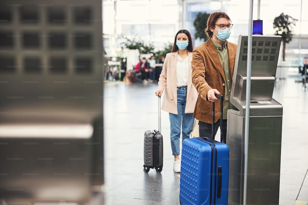 Male traveler in protective face mask checking luggage weight while woman standing in line at safe distance