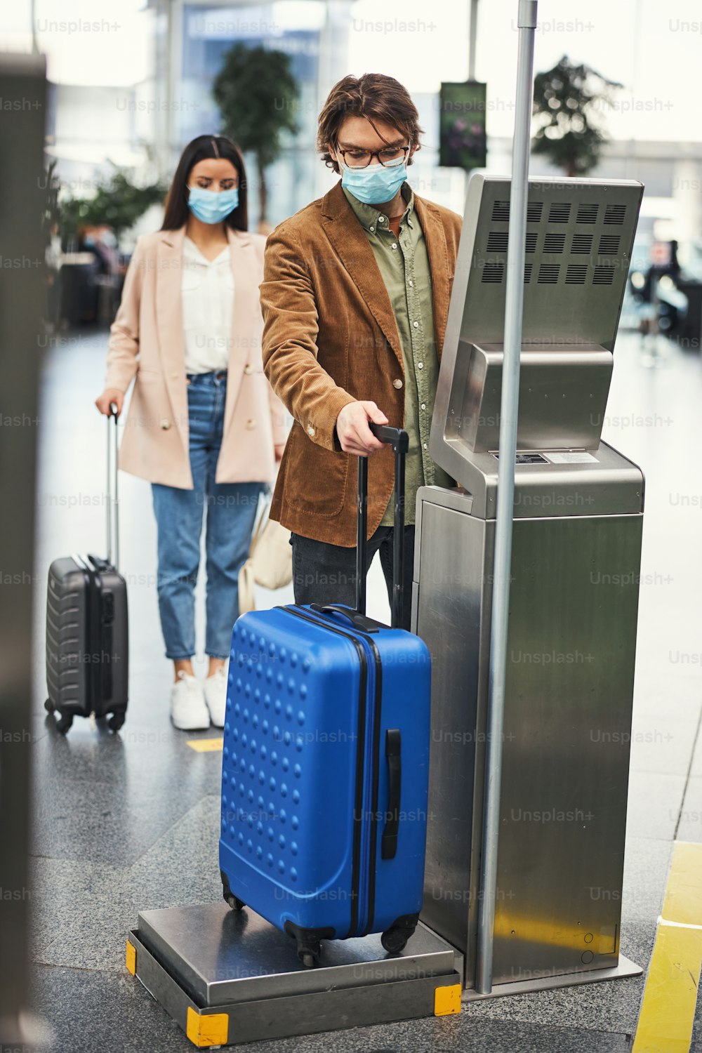 Stylish man in medical mask checking luggage weight while woman standing in line at safe distance