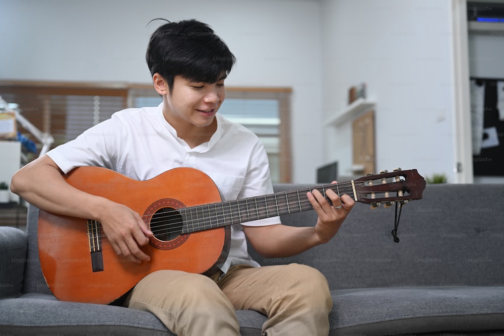 Happy young man playing guitar while siting on comfortable sofa at home.