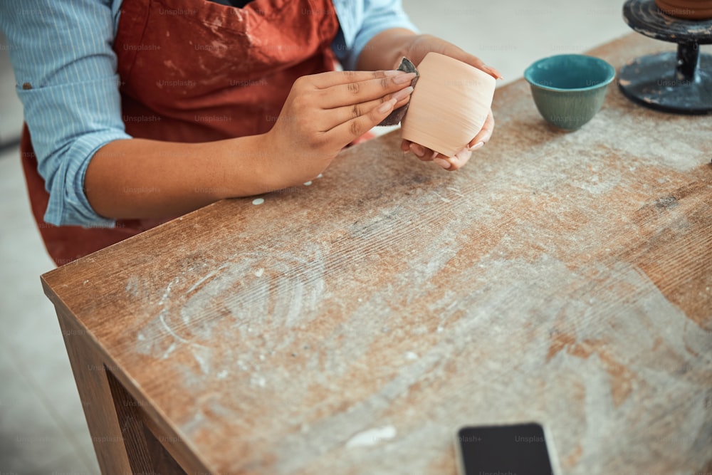 Primer plano de una joven alfarera puliendo y alisando loza con papel de lija mientras está sentada en la mesa en el estudio de cerámica