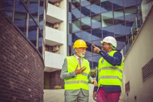 Mature engineer discussing the structure of the building with architects colleague at construction site. Businessmen talking.