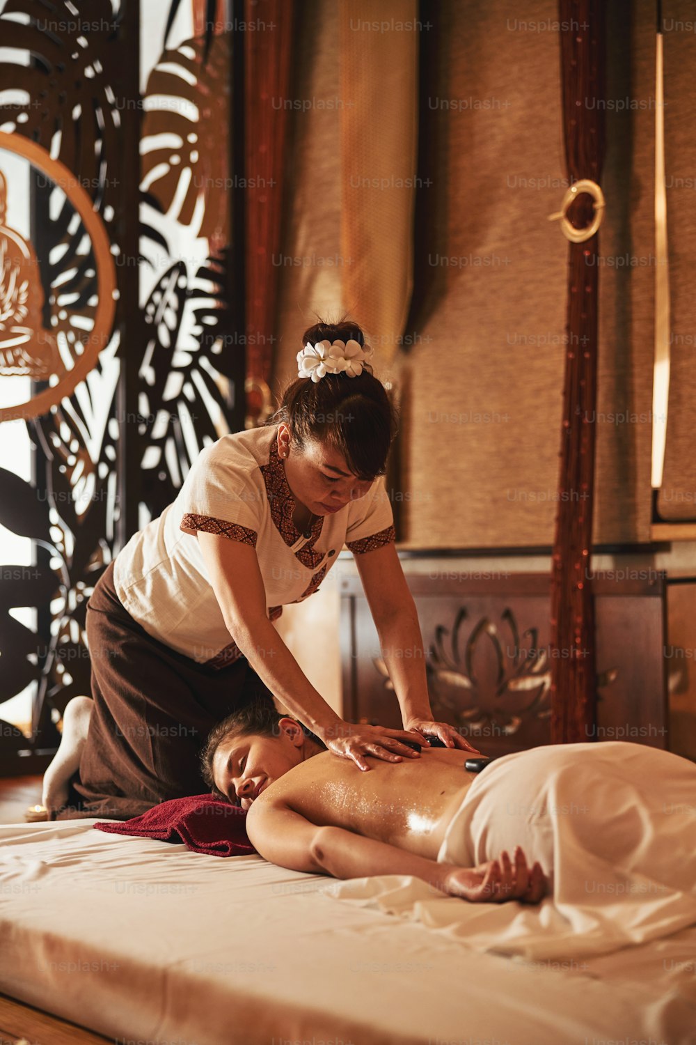 Asiatischer Masseur in einem Raum des Spa-Salons mit heißen Steinen als Werkzeug zur Massage des weiblichen Rückens
