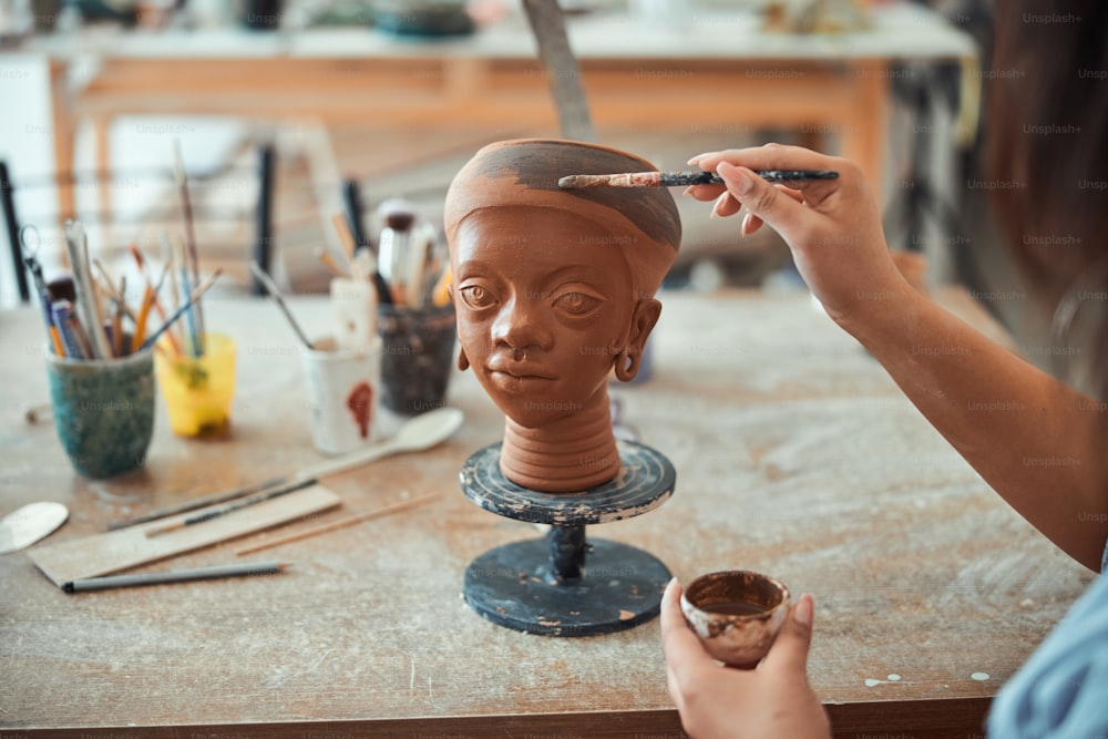 Primer plano de una joven artista de cerámica que pinta un recuerdo de cerámica con un pincel mientras trabaja en un estudio de cerámica artística