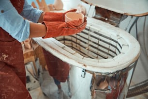電気陶器窯の近くに立っている間、手作りの粘土ボウルを持っている若い女性陶芸家の接写