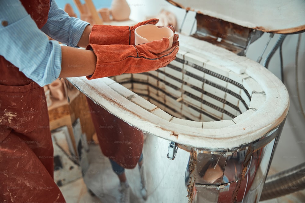 Primer plano de una joven ceramista sosteniendo un cuenco de arcilla hecho a mano mientras está de pie cerca del horno eléctrico de cerámica