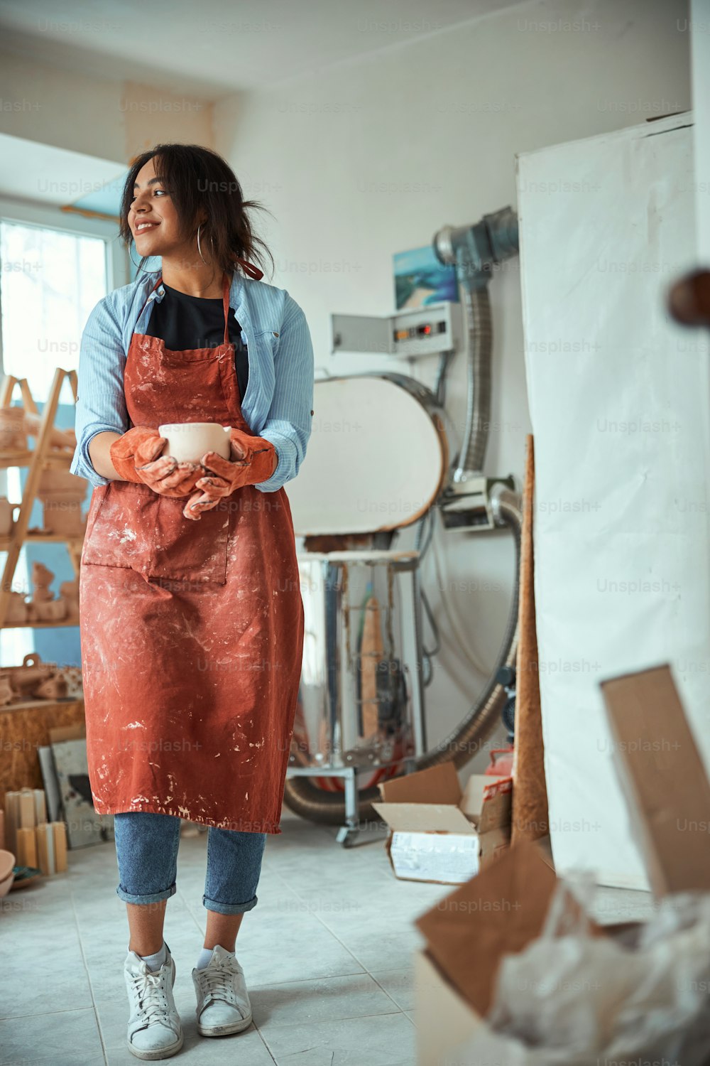 Beautiful young woman ceramic artist holding earthenware bowl and smiling while standing in pottery workshop