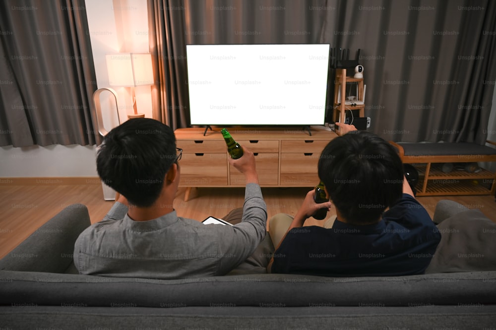 두 명의 아시아 남자가 맥주를 마시고 소파에 앉아 TV를 보고 있다.