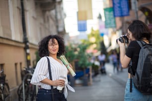 コーヒーを飲みながら通りに立つ笑顔の女性。彼女の写真を撮る女性。焦点は背景にあります。