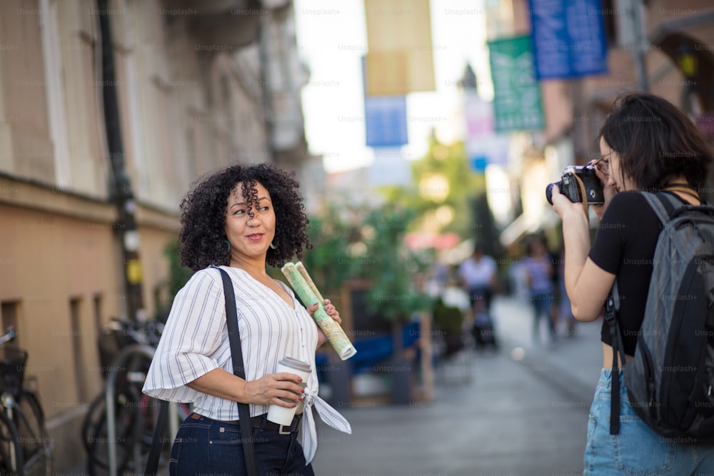 Smiling woman standing on the street with cup of coffee. Woman taking photo of her. Focus is on background.
