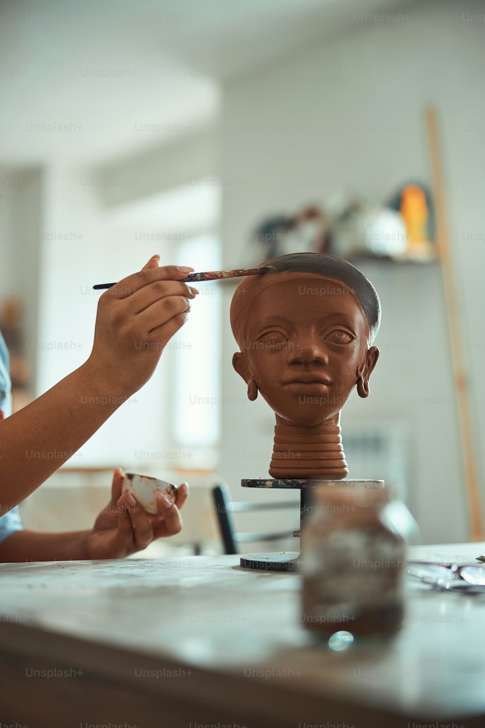 Close up of young woman ceramic artist painting on clay sculpture with paintbrush while working in pottery art studio
