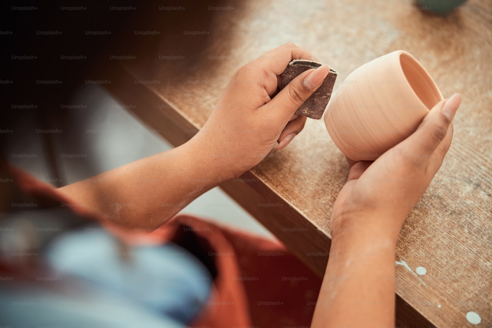 Primer plano de una joven alfarera puliendo cerámica con papel de lija mientras está sentada en la mesa en el estudio de cerámica