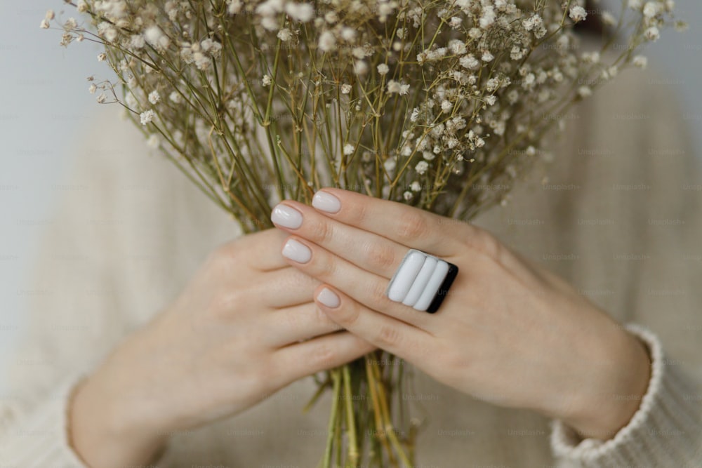 Beautiful stylish woman with modern square ring holding dried flowers, close up. Fashionable female in sweater with unusual fused glass accessories and white manicure. Beauty and care concept