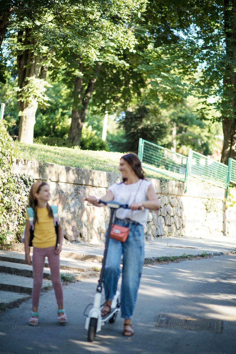 Mother and daughter walking trough park. Woman driving push scooter.