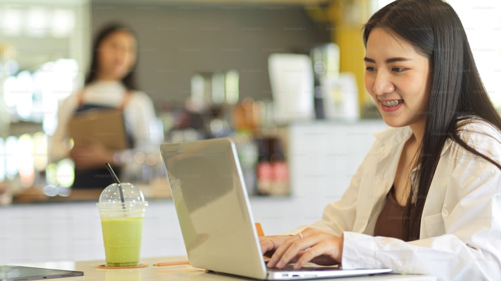 Side view of female teenager smiling and using laptop while sitting in coffee shop