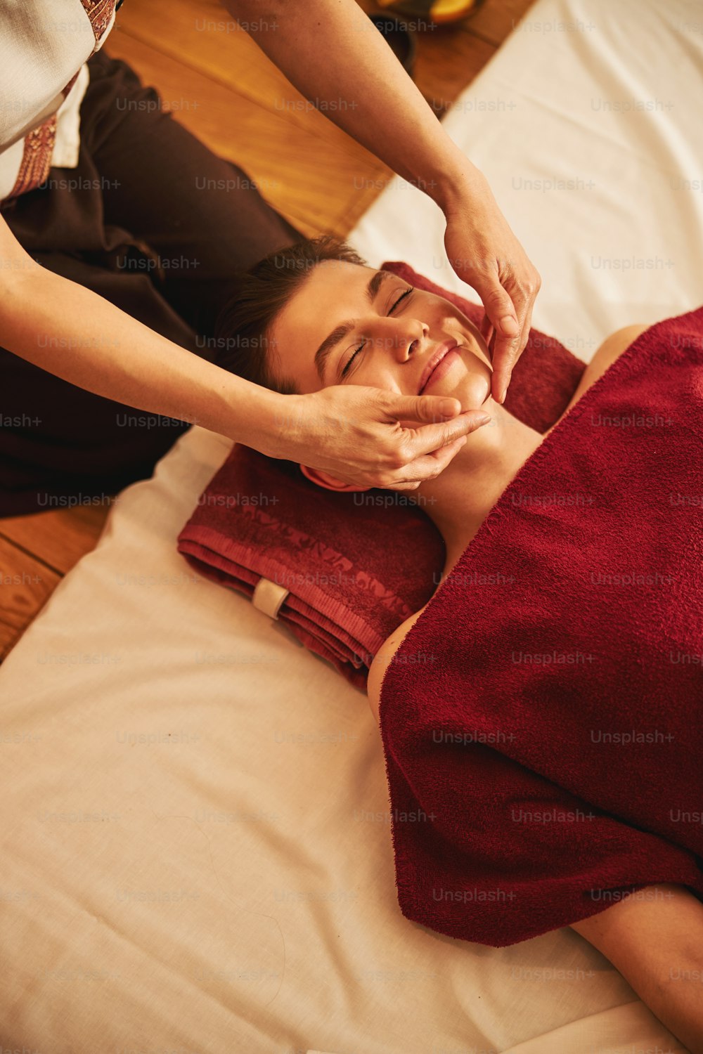 Female with body covered in red blanket enjoying hand movements of massage therapist on sides of her chin