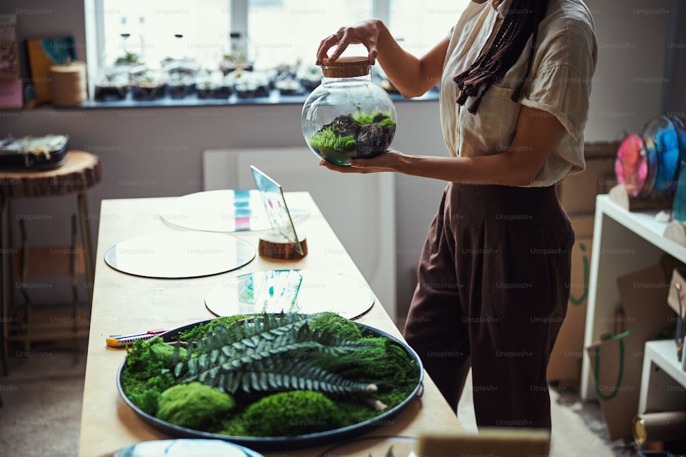 Cropped photo of a certified florist holding a closed round glass jar plant terrarium in front of her