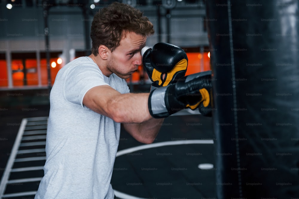 Young man in white shirt and boxing protective gloves doing exercises in gym with pushing bag.