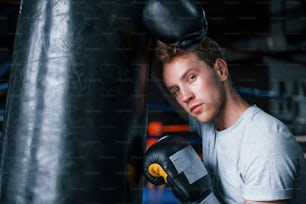 Tired young boxer in white shirt and with protective gloves leaning on punching bag in the gym.