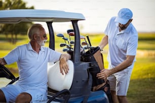 Dois golfistas seniores em quadra. Homem sentado no carrinho de golfe.